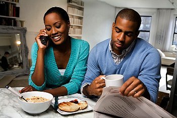 A woman on the phone and a husband reading a paper sitting at their table with breakfast food